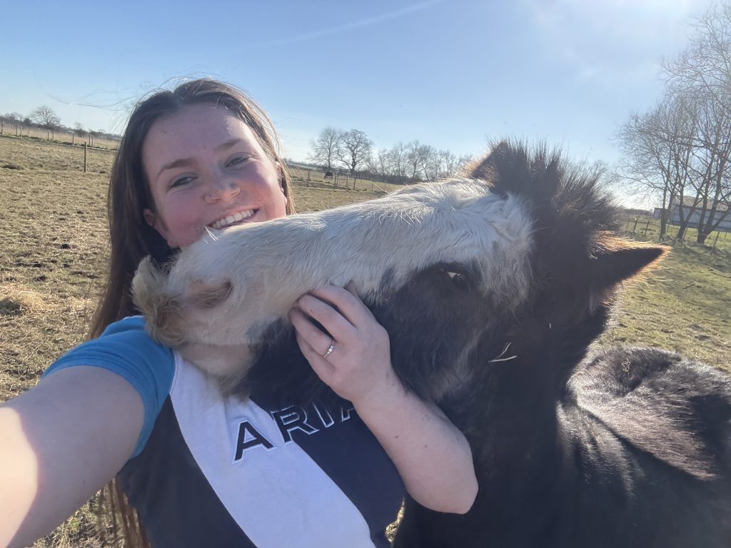 A woman smiles while taking a selfie in a sunny field as a friendly black-and-white pony nuzzles her face affectionately.