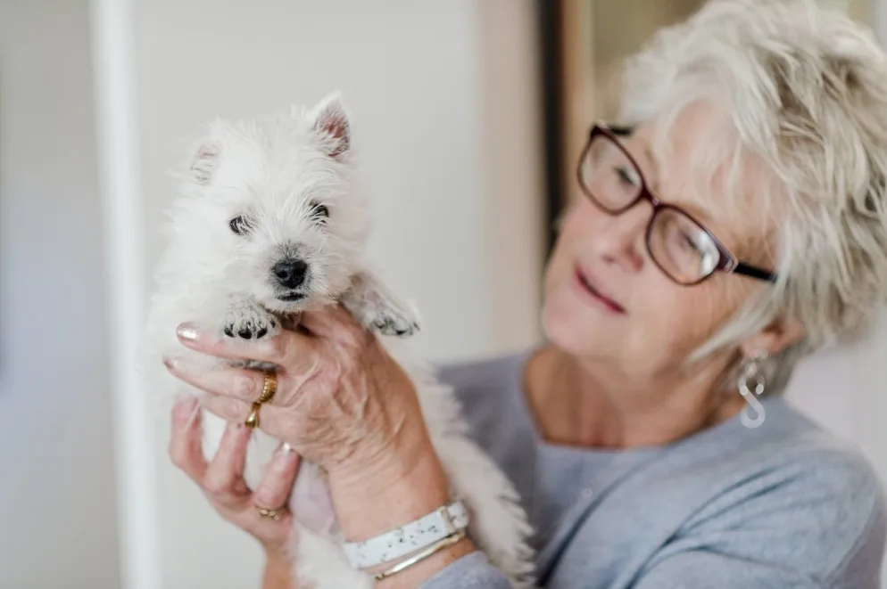 Older woman holding a white rescue puppy in a home environment after adoption